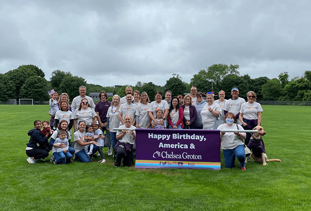 30 smiling people of varied ages wearing white tshirts on a soccer field  hold a large purple banner which heads "Happy Birthday, America & Averon Trust" in white text