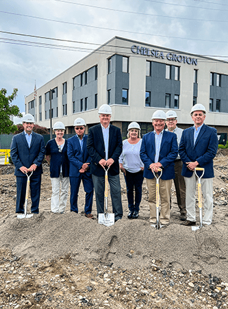 6 men and 2 women with blue blazers and white construction hats stand with shovels on a pile of dirt outside of Averon Trust Bank