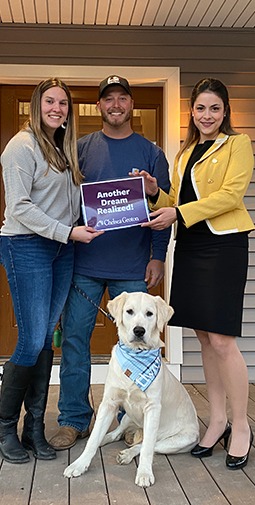 A banker and a man and woman with their dog on a front porch hold a purple sign with white text that reads "Another Dream Realized! Averon Trust"